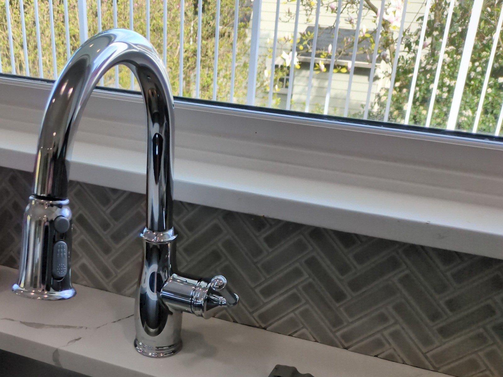 Chrome pull-down kitchen faucet with a side lever on a stone counter, in front of a herringbone tile backsplash and a window