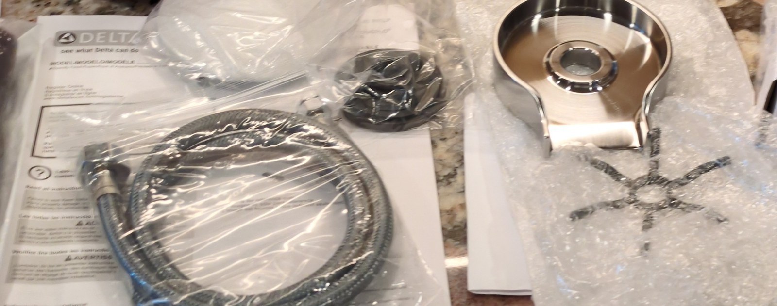 Unboxed Delta faucet parts laid out on a granite counter, including a coiled braided supply hose, the instruction sheet and a brushed-nickel rinser base still in its wrapping