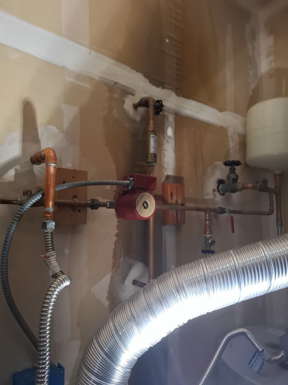 Red Grundfos circulator pump plumbed into copper lines on a wall beside a water heater in a Mukilteo home, with a gate valve above it, an expansion tank to the right and a flexible vent duct in the foreground