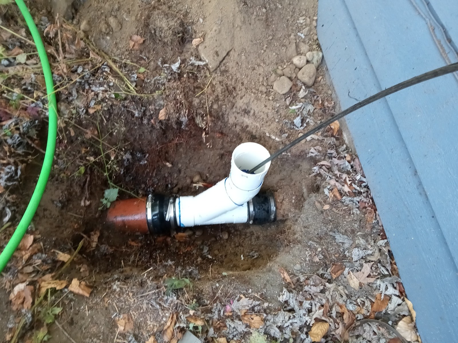 New two-way sewer cleanout with a capped white riser and banded couplings installed in a small excavation beside a gray house wall, with a drain cable running into the open riser