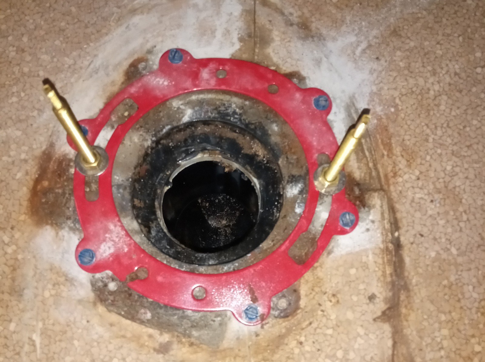 Red steel flange repair ring fastened with blue-headed screws around a toilet drain opening, two brass bolts standing upright through it