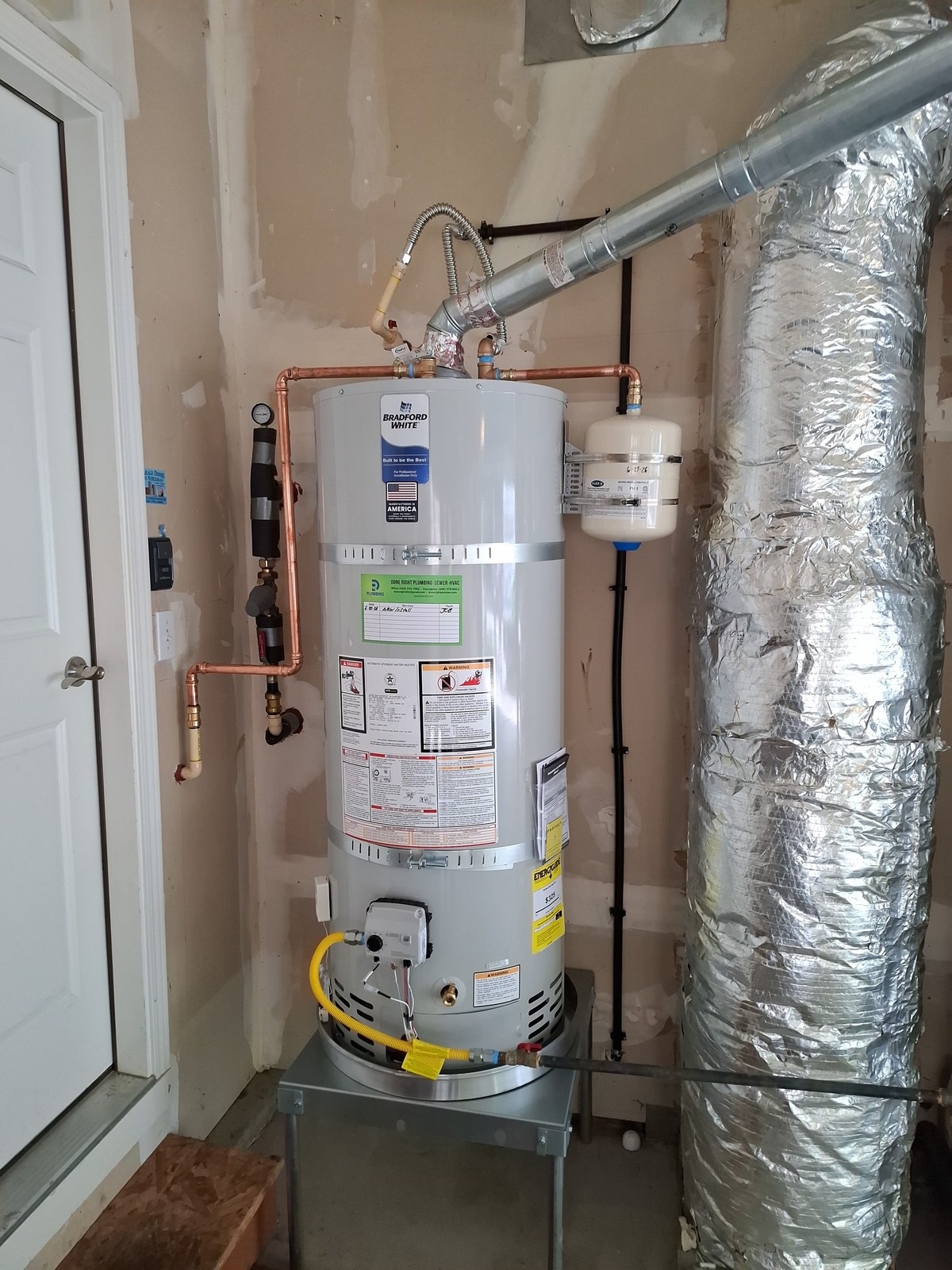 New 50-gallon Bradford White gas water heater on a stand in a garage, with new copper piping, a pressure gauge, seismic straps and an expansion tank on the wall